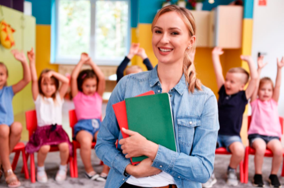 An Educator standing in front of the class room with the children in the back with their hands up