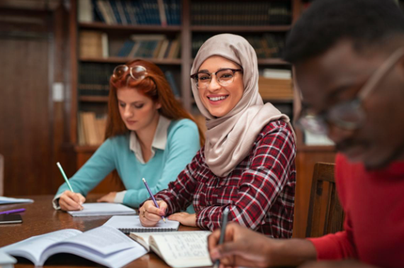 3 adults sitting in a library writing in their books. The middle one is looking at the camera and smiling